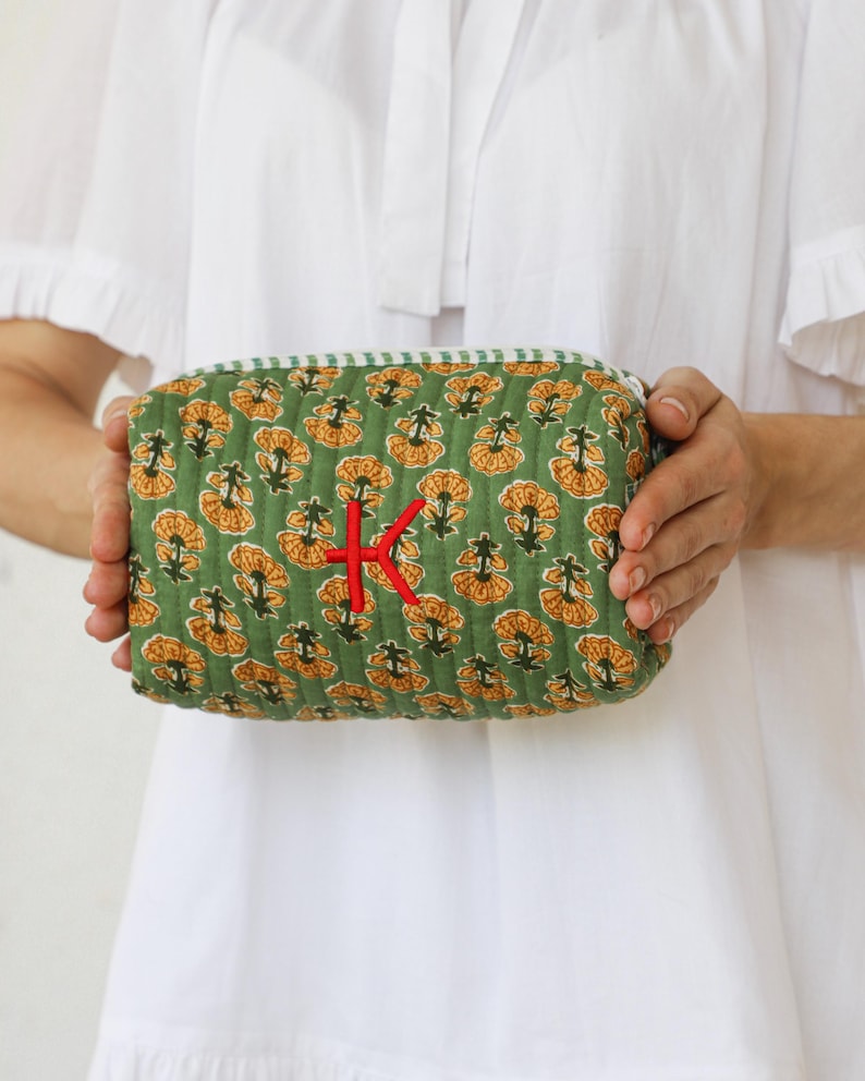 May include: A green quilted cosmetic bag with a floral pattern in yellow and orange. The bag has a red embroidered "K" and a green and white striped trim. The bag is held by a person wearing a white dress.