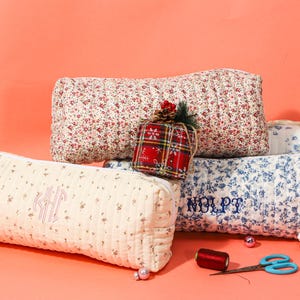 May include: Several quilted cosmetic bags and pillows in floral patterns and a red plaid gift box. The bags are cream, red, and blue with embroidered initials and names. A spool of thread and scissors are also visible.