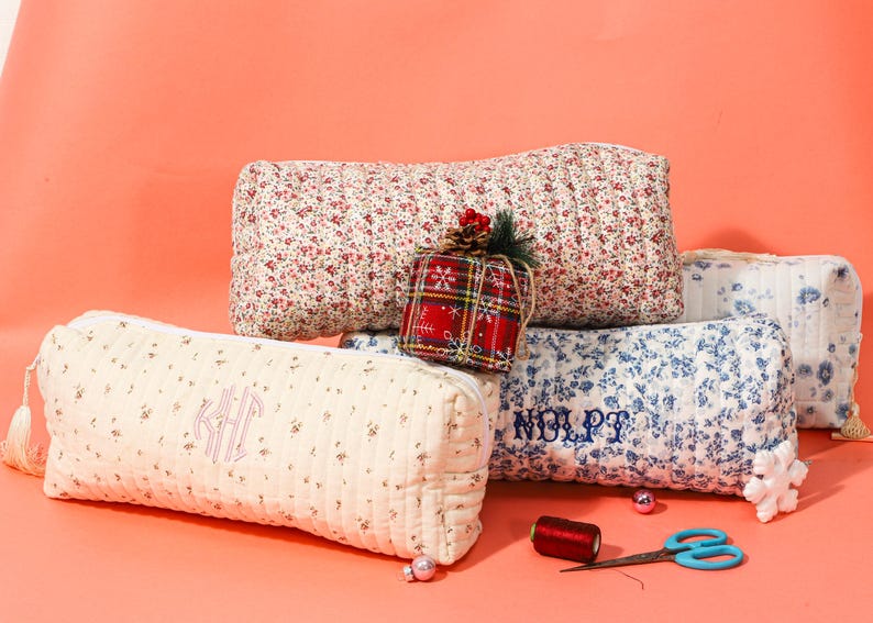 May include: Several quilted cosmetic bags and pillows in floral patterns and a red plaid gift box. The bags are cream, red, and blue with embroidered initials and names. A spool of thread and scissors are also visible.