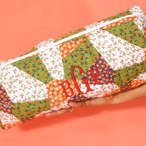 May include: A rectangular quilted cosmetic bag with a white zipper and tassel. The bag features a patchwork design with floral patterns in green, orange, white, and blue. Red embroidered initials are visible on the front.