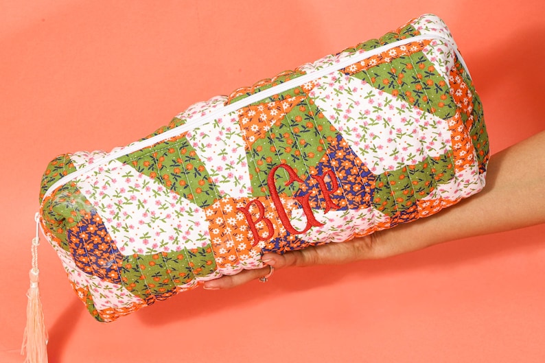 May include: A rectangular quilted cosmetic bag with a white zipper and tassel. The bag features a patchwork design with floral patterns in green, orange, white, and blue. Red embroidered initials are visible on the front.