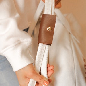 May include: A white canvas tote bag with a brown leather strap and a gold button. The strap has a white cotton cord.