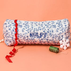 May include: A floral print pillow with blue flowers on a white background, embroidered with the letters "NOLPT" in blue. A red ribbon, a white tassel, a snowflake, and green thread are also present.