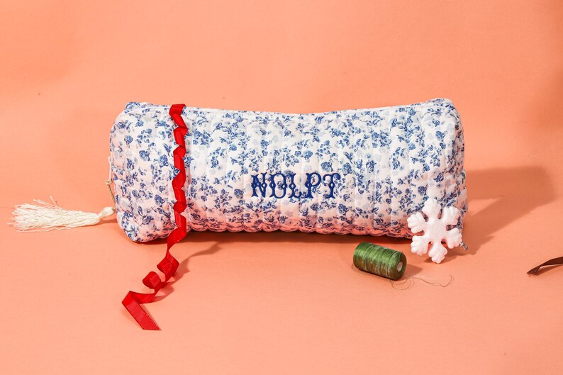 May include: A floral print pillow with blue flowers on a white background, embroidered with the letters "NOLPT" in blue. A red ribbon, a white tassel, a snowflake, and green thread are also present.