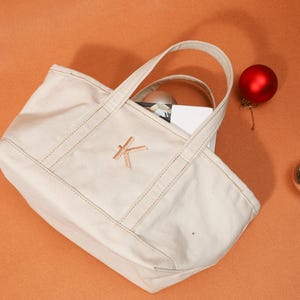May include: A cream canvas tote bag with a stitched 'K' monogram. The bag has two handles and a rectangular shape. A red Christmas bauble and a silver ball sit beside the bag on an orange surface.
