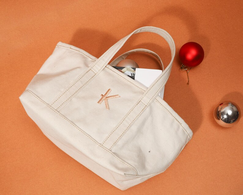 May include: A cream canvas tote bag with a stitched 'K' monogram. The bag has two handles and a rectangular shape. A red Christmas bauble and a silver ball sit beside the bag on an orange surface.