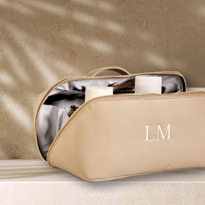 May include: A beige zippered travel makeup bag with a white monogram "LM" on the front. The bag is open and has a white lining. There are two white bottles inside the bag.