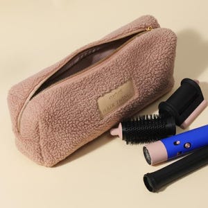May include: A pink, fuzzy zippered pouch with the text "DIOR HAIR THINGS" on a leather patch. Next to it are hair styling tools, including a blue and pink hair styling wand and black attachments.