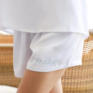Peut inclure: Short blanc avec la date brod&eacute;e "11.11.25" en fil bleu clair. Le short est fait d'un tissu lisse et l&eacute;ger et fait partie d'un ensemble assorti. L'image montre un gros plan du short et d'une partie d'un haut blanc.