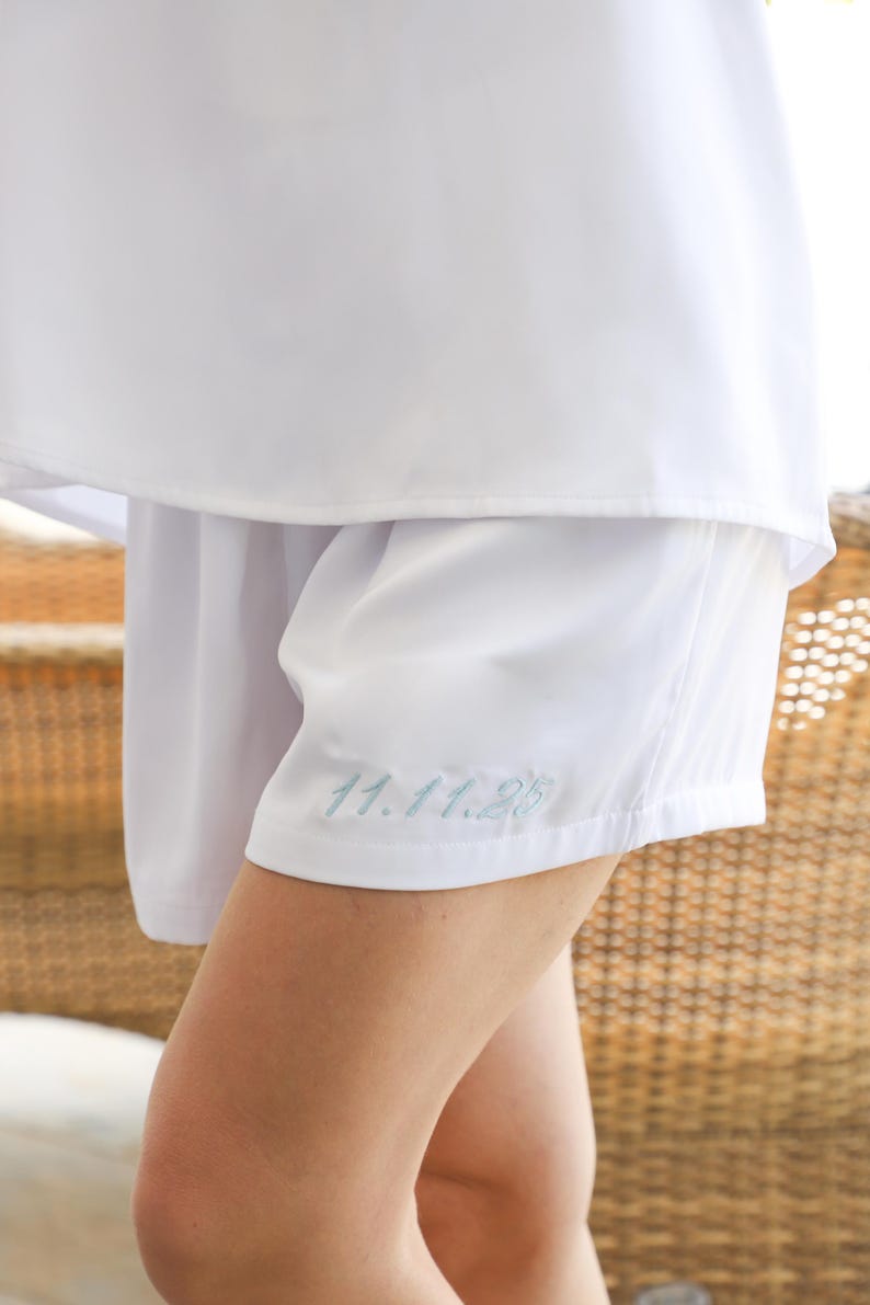 Peut inclure: Short blanc avec la date brod&eacute;e "11.11.25" en fil bleu clair. Le short est fait d'un tissu lisse et l&eacute;ger et fait partie d'un ensemble assorti. L'image montre un gros plan du short et d'une partie d'un haut blanc.