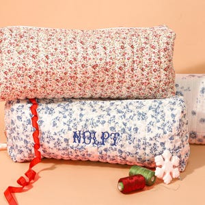 May include: Three floral-patterned quilted pouches in red and white, and blue and white. The blue pouch has embroidered text. A red ribbon, spools of thread, and a snowflake ornament are also present.