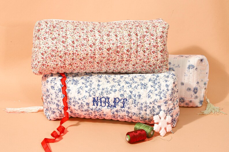 May include: Three floral-patterned quilted pouches in red and white, and blue and white. The blue pouch has embroidered text. A red ribbon, spools of thread, and a snowflake ornament are also present.