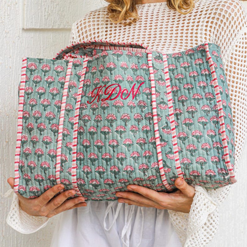Puede incluir: Bolso tote acolchado con un estampado floral en tonos turquesa y rosa. El bolso presenta un ribete y asas a rayas rosas y blancas. El monograma "KDN" est&aacute; bordado en rosa en la parte delantera.