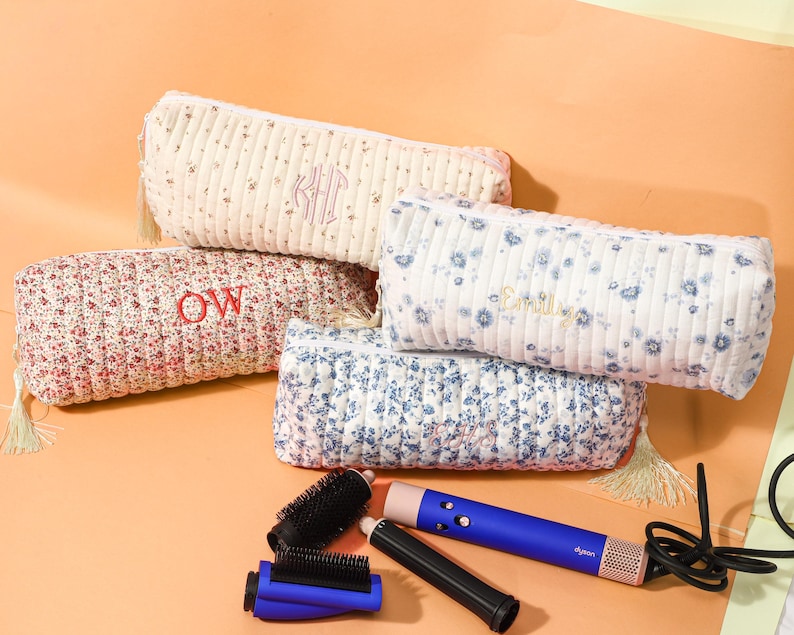 May include: Five floral print cosmetic bags in various colors, with embroidered initials. A blue and pink hair styling tool and accessories are also visible. The bags have tassel zipper pulls.