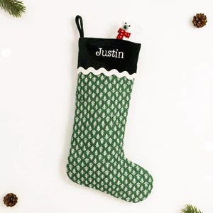 May include: A green and white Christmas stocking with the name "Justin" embroidered in white. The stocking has a black cuff and a white scalloped trim. A small white plush animal with a red scarf peeks out from the top.