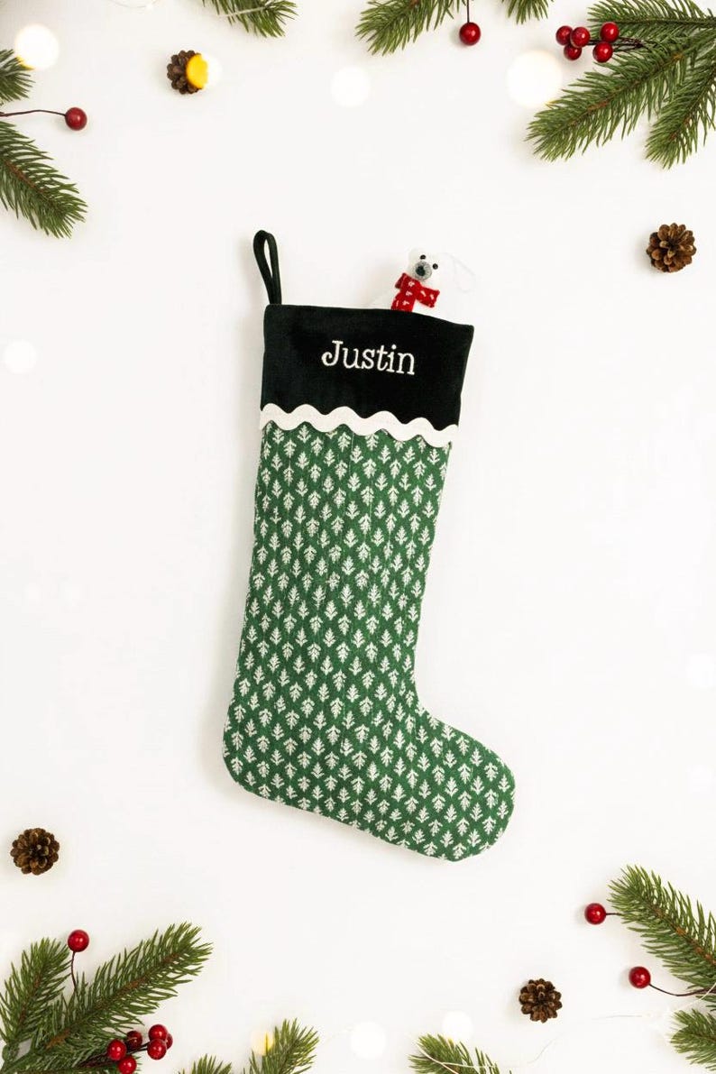 May include: A green and white Christmas stocking with the name "Justin" embroidered in white. The stocking has a black cuff and a white scalloped trim. A small white plush animal with a red scarf peeks out from the top.
