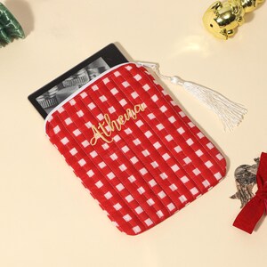 Puede incluir: Funda para lector de libros electr&oacute;nicos a cuadros rojos y blancos con el nombre "Athena" en letras doradas. La funda tiene un tirador de cremallera con borla blanca. Decoraciones navide&ntilde;as rodean la funda, incluido un &aacute;rbol verde y un adorno dorado.