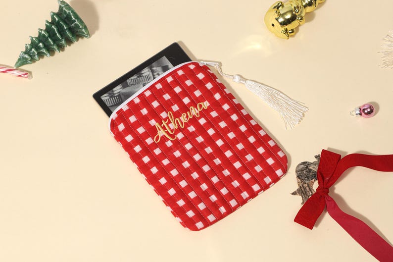 Puede incluir: Funda para lector de libros electr&oacute;nicos a cuadros rojos y blancos con el nombre "Athena" en letras doradas. La funda tiene un tirador de cremallera con borla blanca. Decoraciones navide&ntilde;as rodean la funda, incluido un &aacute;rbol verde y un adorno dorado.