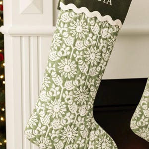 May include: A green and white Christmas stocking with a floral pattern. The stocking has a green cuff with the name "TRISTA" embroidered in white. The stocking is hanging on a white mantel.