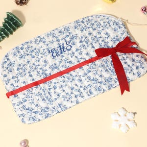 Op de afbeelding: Een gewatteerde make-uptas met een blauw bloemenpatroon en een rood lint. De tas heeft geborduurde initialen en een ritssluiting. Decoratieve elementen zijn een groene kerstboom, een sneeuwvlok en een zuurstok.