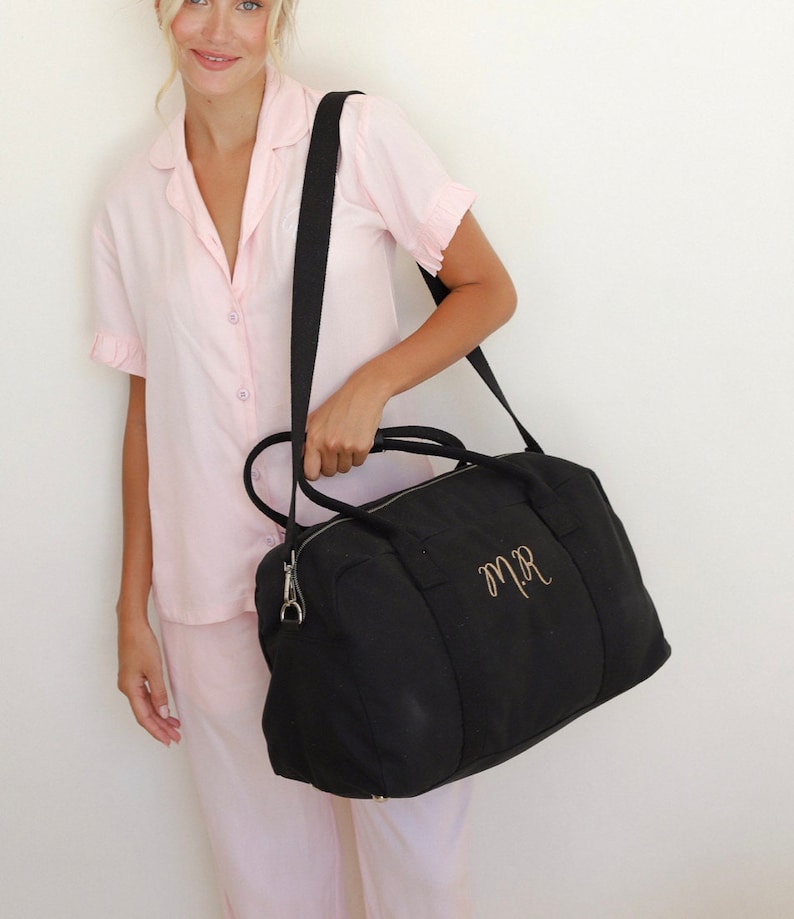 May include: A black canvas travel bag with a gold embroidered monogram "M.R."  The bag has a top zipper closure and a long adjustable shoulder strap.