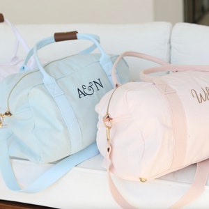 K&ouml;nnte beinhalten: Drei Canvas-Reisetaschen mit personalisierter Stickerei. Die Taschen sind hellblau, rosa und wei&szlig;. Die hellblaue Tasche hat "A&N" gestickt. Die rosafarbene Tasche hat "Willow" gestickt.