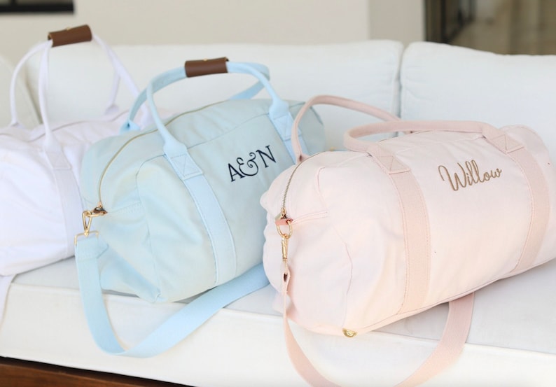 K&ouml;nnte beinhalten: Drei Canvas-Reisetaschen mit personalisierter Stickerei. Die Taschen sind hellblau, rosa und wei&szlig;. Die hellblaue Tasche hat "A&N" gestickt. Die rosafarbene Tasche hat "Willow" gestickt.