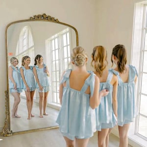 Peut inclure: Plusieurs personnes v&ecirc;tues de pyjamas bleu clair avec des d&eacute;tails en forme de n&oelig;uds se tiennent devant un grand miroir dor&eacute; orn&eacute;. Le miroir refl&egrave;te le groupe, montrant un total de six personnes. La pi&egrave;ce a des murs blancs et de grandes fen&ecirc;tres.