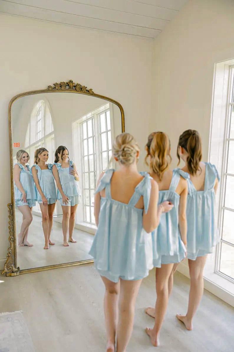 Peut inclure: Plusieurs personnes v&ecirc;tues de pyjamas bleu clair avec des d&eacute;tails en forme de n&oelig;uds se tiennent devant un grand miroir dor&eacute; orn&eacute;. Le miroir refl&egrave;te le groupe, montrant un total de six personnes. La pi&egrave;ce a des murs blancs et de grandes fen&ecirc;tres.