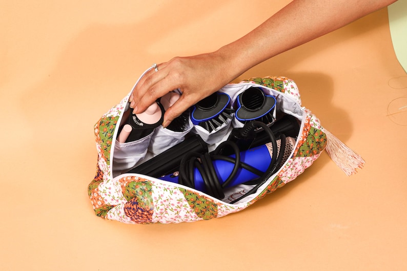 May include: A floral patterned cosmetic bag filled with hair styling tools. The bag contains a black and blue hair dryer, brushes, and other styling accessories. The bag is open, and a hand is reaching inside.