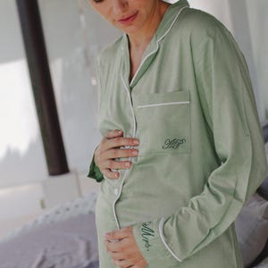 May include: A sage green, long-sleeved button-down shirt with white piping. The shirt has a pocket on the left chest with the initials "HP" embroidered in black. The words "Mrs." are embroidered on the left cuff in black.