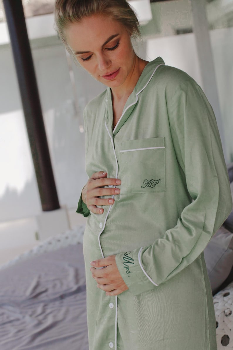 May include: A sage green, long-sleeved button-down shirt with white piping. The shirt has a pocket on the left chest with the initials "HP" embroidered in black. The words "Mrs." are embroidered on the left cuff in black.