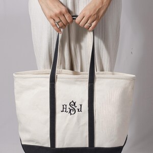 May include: Cream-colored canvas tote bag with black accents and handles. The bag features embroidered initials in black. The bottom and handles are black. The bag is held by a person wearing rings.