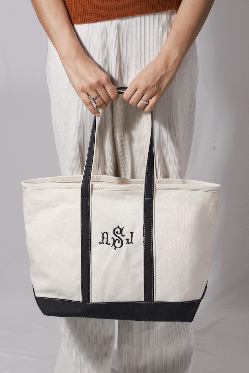 May include: Cream-colored canvas tote bag with black accents and handles. The bag features embroidered initials in black. The bottom and handles are black. The bag is held by a person wearing rings.