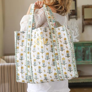 Puede incluir: Bolso tote blanco con un estampado floral amarillo y ribete a rayas verdes. El bolso tiene una textura acolchada y un monograma amarillo. Las asas tambi&eacute;n tienen flores amarillas y rayas verdes. Un accesorio elegante y pr&aacute;ctico.
