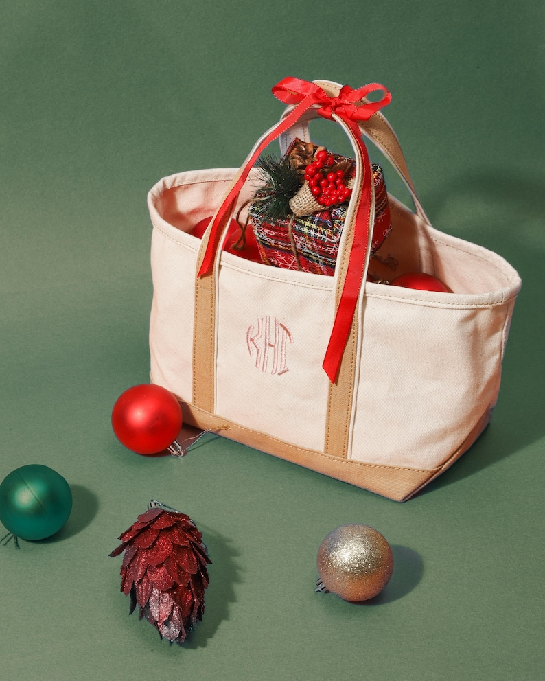 May include: A canvas tote bag with a monogram, tan trim, and red ribbon. The bag is filled with Christmas ornaments and a wrapped gift. A red, green, and gold ornament are scattered around the bag on a green background.