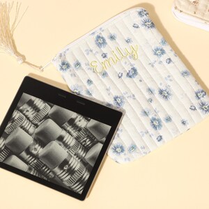 Puede incluir: Un lector electr&oacute;nico con pantalla negra que muestra un patr&oacute;n de l&aacute;pices, junto a una bolsa acolchada blanca con estampado floral azul y el nombre "Emily" bordado en amarillo. Una borla cuelga de la bolsa.