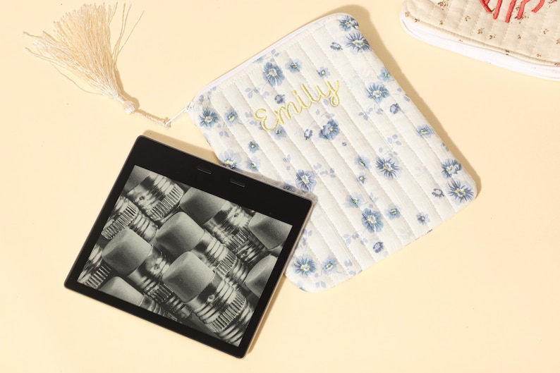 Puede incluir: Un lector electr&oacute;nico con pantalla negra que muestra un patr&oacute;n de l&aacute;pices, junto a una bolsa acolchada blanca con estampado floral azul y el nombre "Emily" bordado en amarillo. Una borla cuelga de la bolsa.