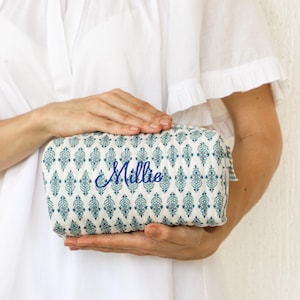 May include: A rectangular cosmetic bag with a blue and white patterned design. The bag has a zipper closure and the name "Millie" embroidered in blue script. The bag is held by a person wearing a white dress.
