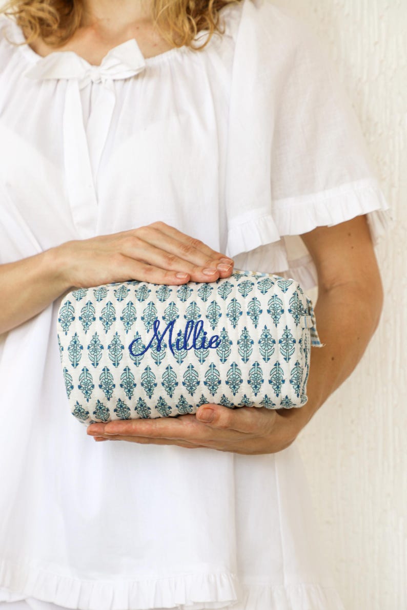 May include: A rectangular cosmetic bag with a blue and white patterned design. The bag has a zipper closure and the name "Millie" embroidered in blue script. The bag is held by a person wearing a white dress.