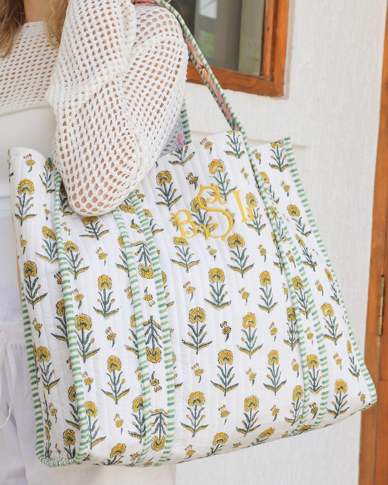 Puede incluir: Bolso tote acolchado blanco con un estampado floral amarillo y ribete a rayas verdes. El bolso presenta iniciales bordadas en amarillo y una correa estampada. Dise&ntilde;ado para transportar objetos.