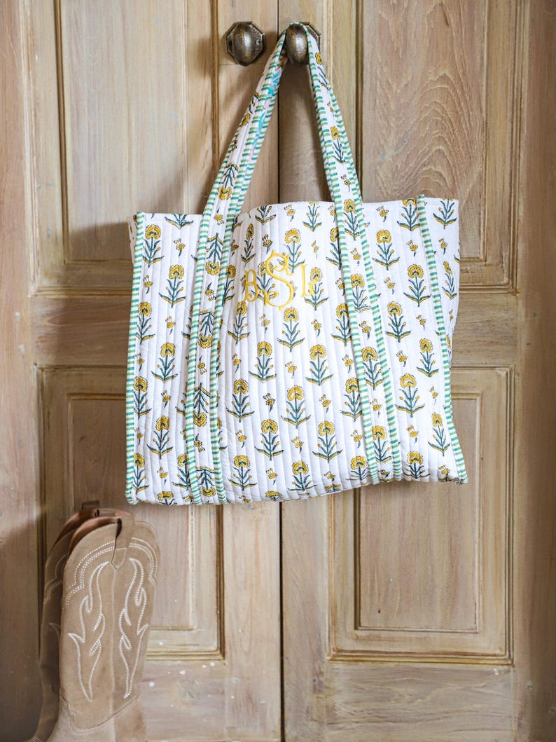 Puede incluir: Un bolso tote blanco con un estampado floral amarillo y ribetes verdes cuelga de un tirador de puerta. La bolsa presenta un dise&ntilde;o acolchado y un monograma amarillo. Un par de botas de vaquero color canela son visibles debajo.