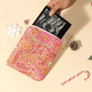 Puede incluir: Una funda rosa para tableta con estampado floral y interior blanco, que contiene un lector electr&oacute;nico negro. La funda presenta un dise&ntilde;o acolchado con elementos florales naranjas y verdes. Elementos decorativos: copo de nieve, adorno, pi&ntilde;a y bast&oacute;n de caramelo.
