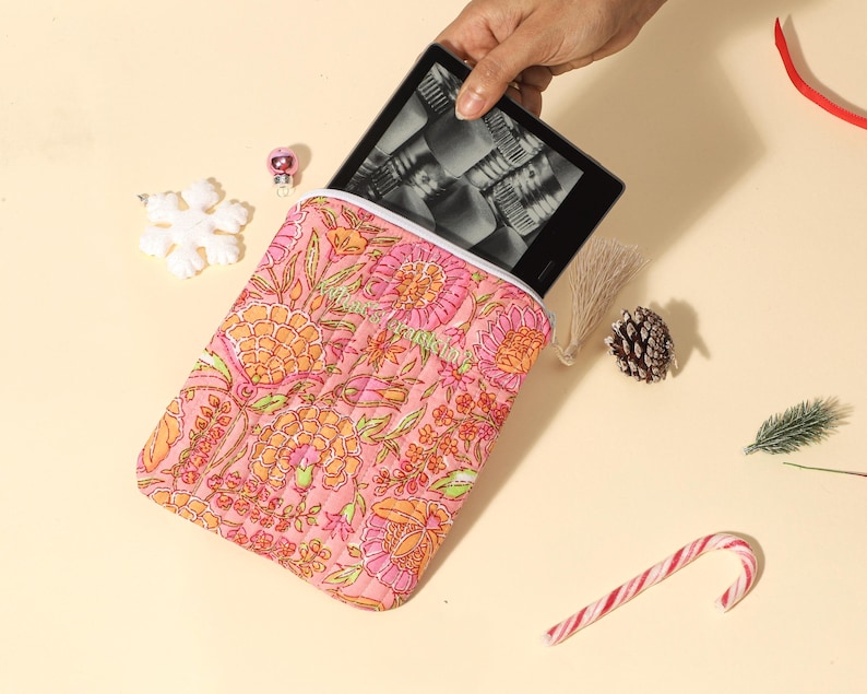 Puede incluir: Una funda rosa para tableta con estampado floral y interior blanco, que contiene un lector electr&oacute;nico negro. La funda presenta un dise&ntilde;o acolchado con elementos florales naranjas y verdes. Elementos decorativos: copo de nieve, adorno, pi&ntilde;a y bast&oacute;n de caramelo.