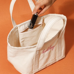 May include: A cream canvas tote bag with the pink embroidered text "book bag". A black lipstick tube with a silver cap and the "Dior" logo is held inside the bag's pocket. The bag has two handles and a zipped pocket.