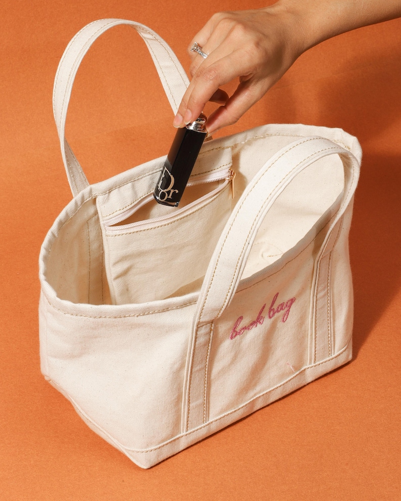 May include: A cream canvas tote bag with the pink embroidered text "book bag". A black lipstick tube with a silver cap and the "Dior" logo is held inside the bag's pocket. The bag has two handles and a zipped pocket.