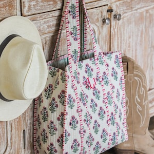 Puede incluir: Un bolso tote blanco con un estampado floral en rosa y azul cuelga de un armario de madera. El bolso tiene un ribete y asas rojas. Tambi&eacute;n se ven un sombrero de paja y un par de botas de vaquero.
