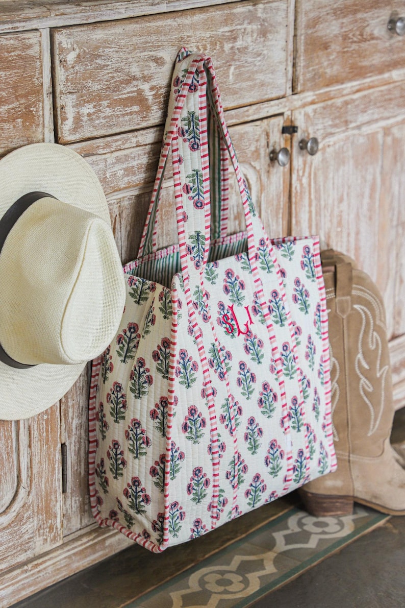 Puede incluir: Un bolso tote blanco con un estampado floral en rosa y azul cuelga de un armario de madera. El bolso tiene un ribete y asas rojas. Tambi&eacute;n se ven un sombrero de paja y un par de botas de vaquero.