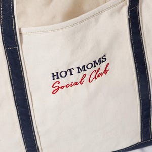 May include: Cream-colored tote bag with navy blue trim and handles. The words "HOT MOMS Social Club" are embroidered in navy and red thread on the front. The bag appears to be made of a sturdy canvas material.