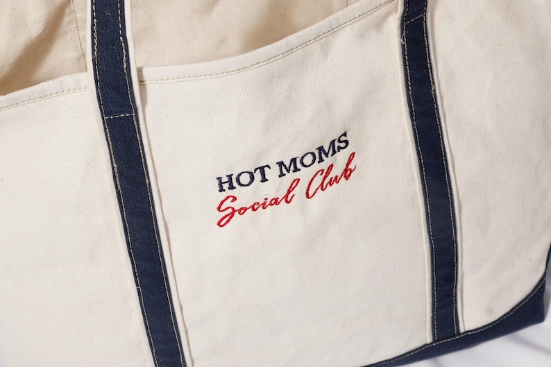 May include: Cream-colored tote bag with navy blue trim and handles. The words "HOT MOMS Social Club" are embroidered in navy and red thread on the front. The bag appears to be made of a sturdy canvas material.